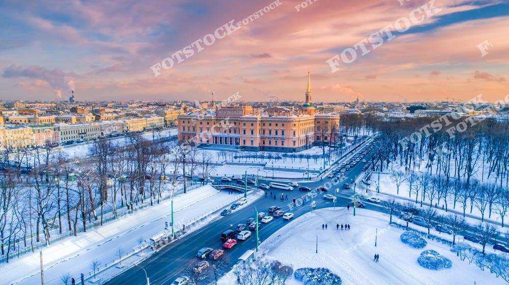 Панорамный зимний вид на Санкт-Петербург с высоты во время яркого заката, открывающий исторический центр города и знаменитый Михайловский замок, также известный как Инженерный замок. Заснеженные улицы, архитектура старого Петербурга и тёплые оттенки закатного неба создают атмосферный городской пейзаж северной столицы России. Фотография подчёркивает величие исторической архитектуры, уникальный стиль городских улиц и романтику зимнего Петербурга. Изображение идеально подходит для публикаций о путешествиях по России, архитектуре Санкт-Петербурга, зимнем туризме, городских панорамах и достопримечательностях России.